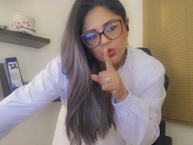 Sofias, con cabello castaño largo, gafas y camisa blanca, haciendo un gesto de silencio con el dedo en los labios, llevando un anillo, con expresión seria, en una habitación con persianas de madera..