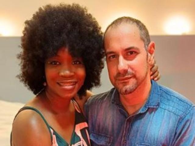 MaxAndMegan69 with big black afro and green top, next to bald MaxAndMegan69 with blue shirt, arm around her, both smiling. Warm light in the background.