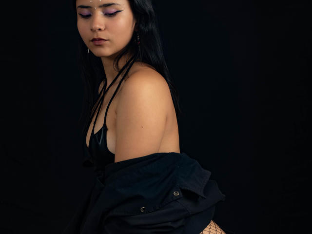 YanNyan, avec ses cheveux noirs, son eye-liner violet et son rouge à lèvres foncé, porte un haut noir et un gilet, laissant apparaître ses épaules nues et une peau légèrement bronzée, sur un fond sombre..