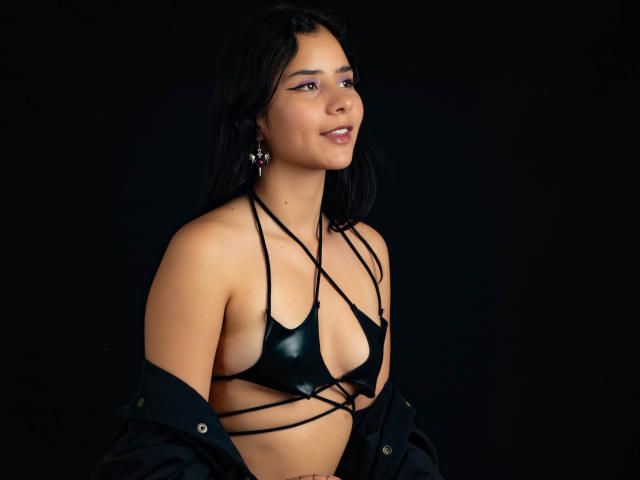 YanNyan with black shiny bikini, strappy top, showing her small tits, black jacket off, dark hair, earrings, smiling, dark background.