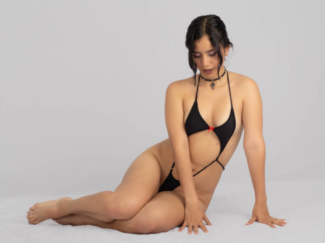 YanNyan, wearing a black bikini with red accent, sits on the floor, legs crossed, arm behind, looking down, showing her tits and pussy, with a black choker.