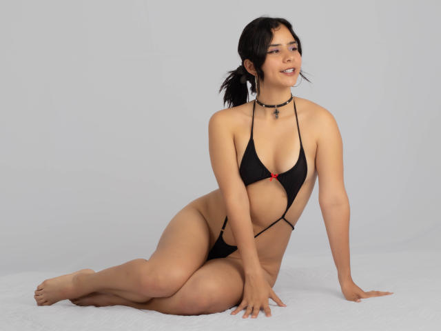 YanNyan in a black string bikini with a small red bow, sitting on the floor, legs folded, arms behind, looking at the camera, wearing a black choker.
