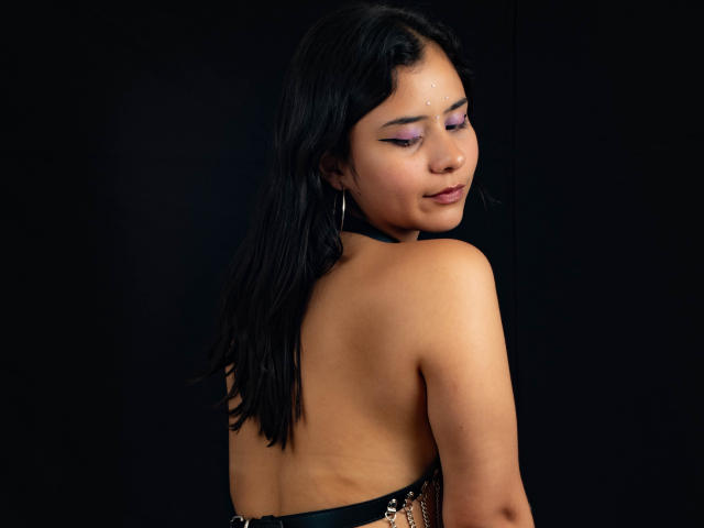 YanNyan with dark hair, purple eye makeup, and a small dot on her forehead, topless, showing her back and ass, looking over her shoulder, black background.