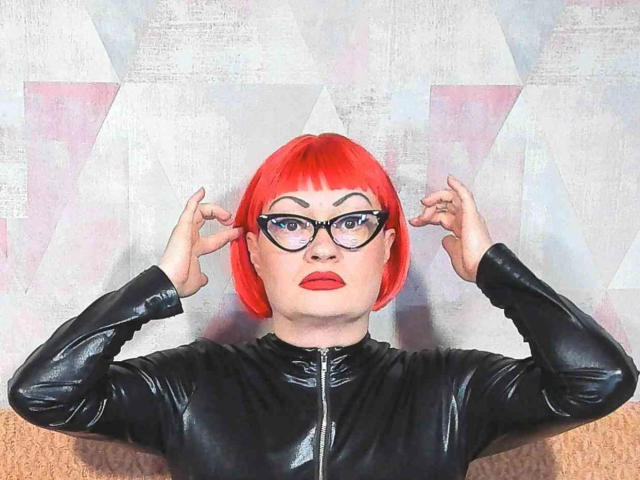 Lackengel-hot with bright red bob, black glasses, and red lipstick, wearing a shiny black latex top, arms raised, fingers touching ears, staring confidently.