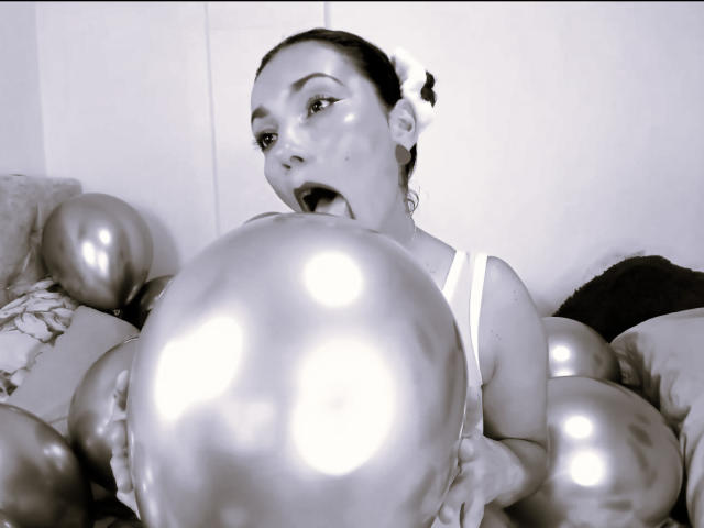 AmberDemons with shiny balloons in her mouth, tongue out, wearing a white tank top, looking up with a surprised expression, holding a large, metallic balloon. Black and white.