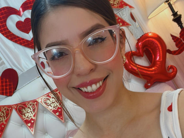 NatyBakers con rossetto rosso, occhiali trasparenti e una camicetta bianca, sorride alla fotocamera, mostrando i denti. In secondo piano, palloncini a forma di cuore rossi e festoni..