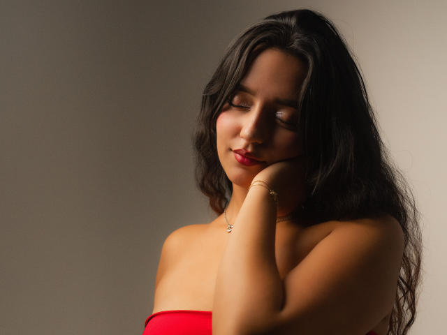 MelissaSmithers with dark, wavy hair, red lipstick, and closed eyes, resting her chin on her hand, wearing a red strapless top, and a gold bracelet, smiling softly.
