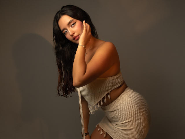 MelissaSmithers with long black hair, tan skin, and a curvy ass, wearing a beige, off-shoulder, fringed knitted top and skirt, leaning forward, hand on cheek, looking at camera.
