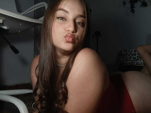 TaniaCooper69 with long brown hair, pouty lips, green eyes, wearing a red top, lying on floor, white chair in background, showing her bare ass, necklace, and relaxed expression.