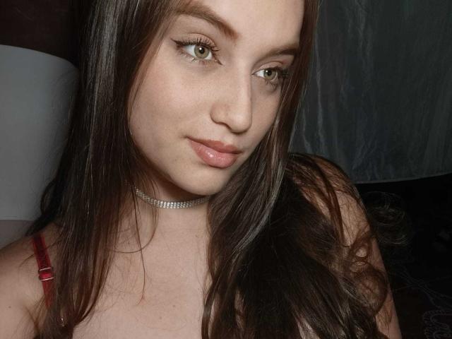 TaniaCooper69 with long brown hair, light skin, green eyes, and pink lips, wearing a red strap and a sparkly choker, looking at the camera with a slight smile.
