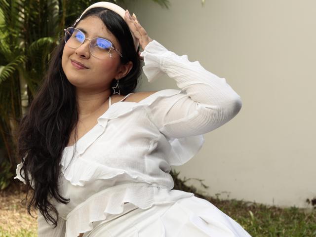 TereCarpert con cabello largo y negro, vestida con un vestido blanco con volantes, gafas azules reflectantes y una diadema blanca, sonriendo, con la mano en el cabello, aretes verdes, fondo de pared blanca..