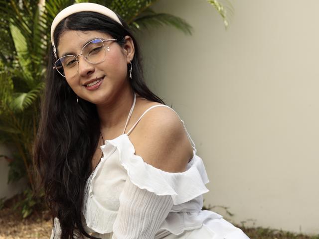 TereCarpert has long black hair, glasses, and a white off-shoulder dress. She's smiling with a cute face, wearing a beige hat, and has small earrings.