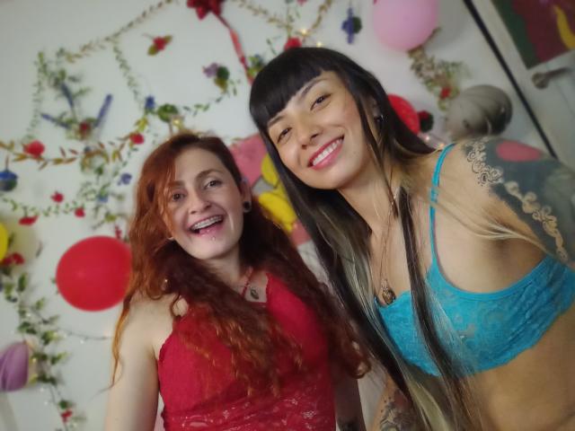 BethAndRoseI con i capelli rossi, con il seno scoperto in una maglia rossa in pizzo, sorridente. BethAndRoseI con i capelli neri, reggiseno blu in pizzo, tatuaggi, sorridente. Entrambe hanno le labbra rosa, sfondo bianco..
