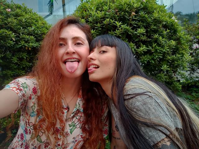 BethAndRoseI with curly red hair and tongue out, and BethAndRoseI with black hair and tongue out, standing in front of green bushes, both looking at the camera.