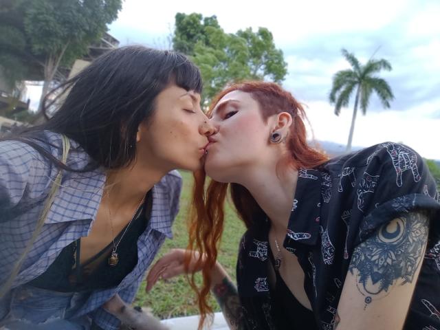 BethAndRoseI with black hair and blue check shirt kissing BethAndRoseI with red hair and black patterned shirt. Red-haired BethAndRoseI has a tattoo on her right arm.