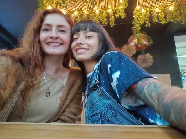 BethAndRoseI, con i capelli rossi e ricci e un seno di dimensioni medie, indossa un cardigan marrone, e Rose, con il caschetto nero e i tatuaggi, indossa una camicia blu con un motivo a stelle, sorridendo..