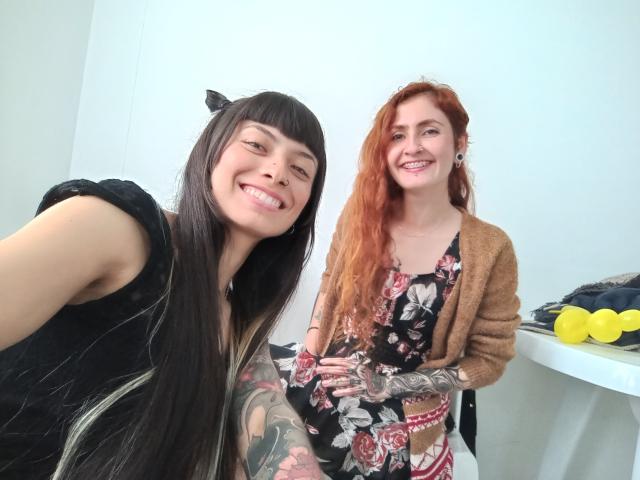 BethAndRoseI con cabello negro largo y una blusa negra, sonriendo a la cámara. RoseAndBethI con cabello rojo, vestido floral y brazos tatuados, vistiendo un cárdigan marrón..