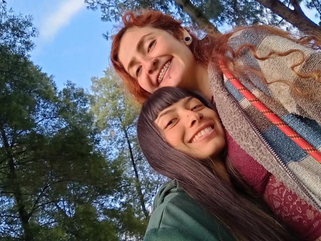 BethAndRoseI, con cabello rizado rojo y piercings en las orejas, sonriendo, tiene los senos cubiertos por un suéter estampado, y está de pie detrás de BethAndRoseII, que tiene cabello lacio y oscuro, una sudadera verde y también está sonriendo..