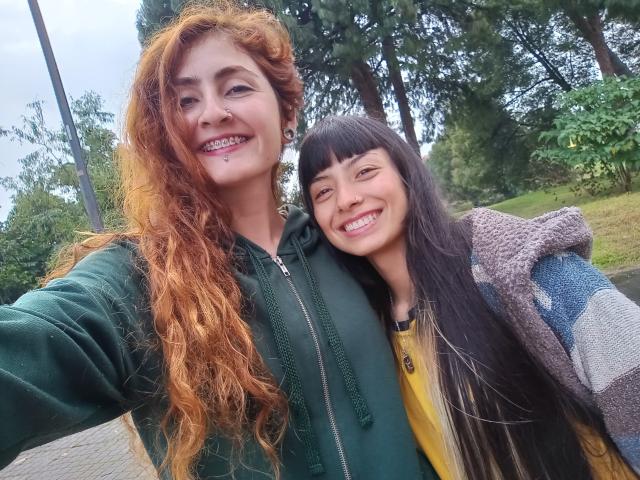 BethAndRoseI con capelli rossi ricci e piercing al naso, felpa verde, e BethAndRoseI con capelli neri lisci, frangia, maglietta gialla e giacca blu a fantasia, sorridenti all'aperto..