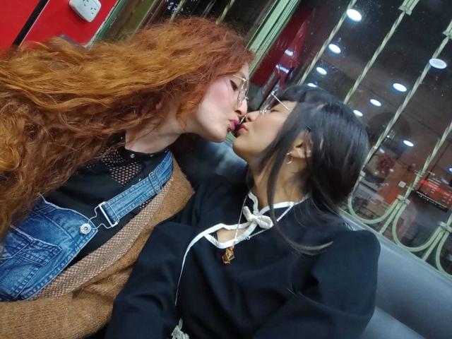 BethAndRoseI, con i capelli rossi ricci, sta baciando BethAndRoseII, con i capelli neri. BethAndRoseII ha dei seni piccoli, indossa una maglietta nera con bordi bianchi, mentre BethAndRoseI indossa una tuta in denim..