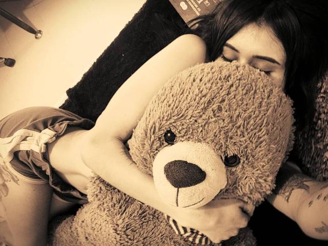 DarkBabe with tattoos, hugging a big fluffy brown teddy bear, nose buried in its nose, wearing loose shorts, her ass slightly visible, black hair, sepia tone.