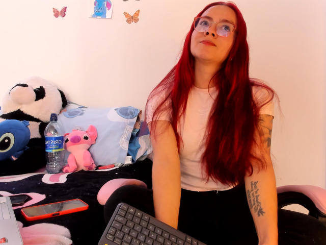 EimmyHoskX with red hair, glasses, white t-shirt, black pants, tattoos, sitting at desk with stuffed toys, water bottle, keyboard, and phone.