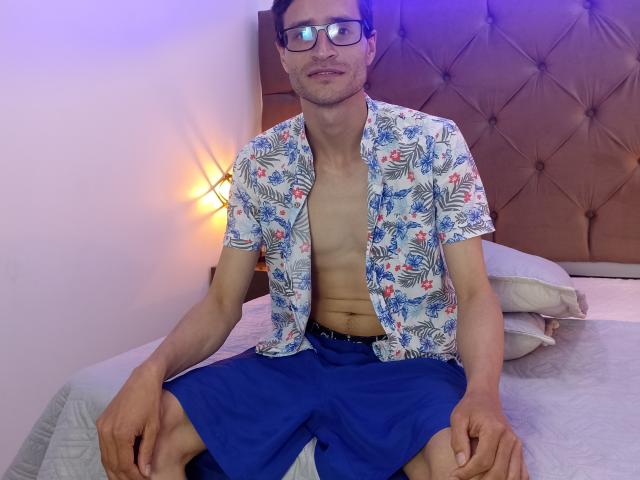 JackkFox, met een open shirt, waardoor een slanke, blote borst zichtbaar is, draagt een blauw bloesjem en blauwe korte broek, zit op een bed, draagt een bril, heeft kort bruin haar, een lichte glimlach, en de slaapkamer heeft een gestoffeerd hoofdeinde..
