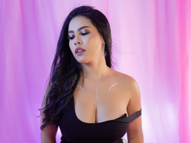 PaolaYKatty with black hair, wearing a black off-shoulder top, showing her big tits, adorned with sparkling jewelry, looking down with a sultry expression. Pink and purple lighting.