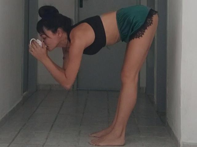 StormArella bends over, drinking from a white cup, wearing a black sports bra and green shorts with black lace, barefoot on gray tiled floor, in a narrow hallway.