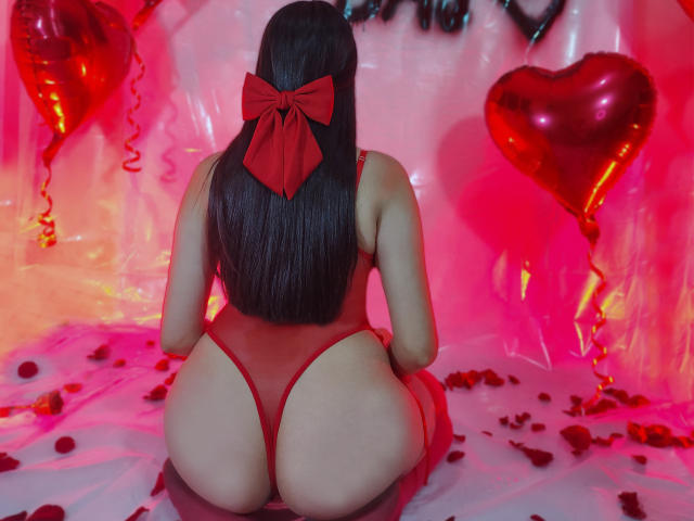 KylieAndJustin is kneeling, facing away, wearing a red thong that reveals her big ass and a red bow in her long black hair. Red heart balloons and petals surround.