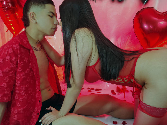 KylieAndJustin, with long black hair, in red lingerie and garter, leans toward Justin, shirtless in red floral shirt, kissing her nose. Red heart balloons and petals on floor.