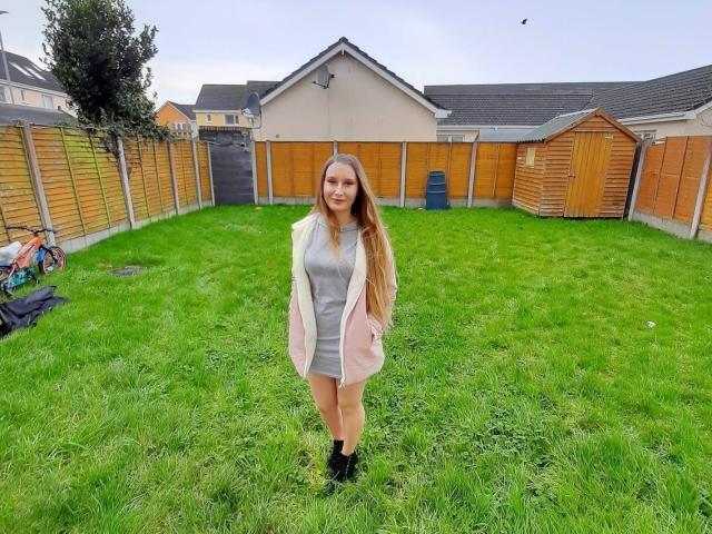 RoxanneStar69 ha lunghi capelli castani, indossa una giacca rosa sopra un vestito grigio, stivali neri, ed è in piedi in un cortile con erba verde, sorridendo, con recinzioni di legno e case sullo sfondo..