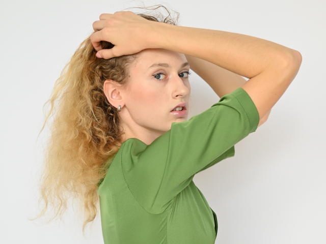 RinaPinkThree with curly blonde hair, green shirt, arms raised, hands in hair, looking to side, subtle makeup, small earrings, white background.