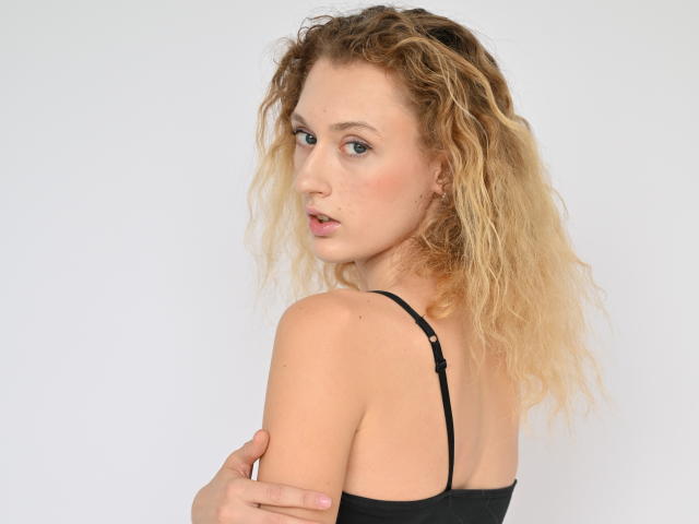 RinaPinkThree with curly blonde hair, blue eyes, and light skin, wearing a black strap top, looking over her shoulder, arms crossed. Boobs visible, subtle makeup.