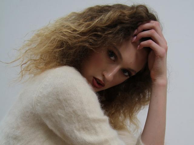 RinaPinkThree with curly blonde hair, wearing a white sweater, looking up with a slightly open mouth, hand in her hair, showing her tits, against a plain background.