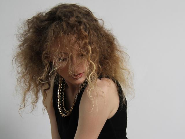 RinaPinkThree has messy, curly brown hair covering her eyes, wearing a black sleeveless top with a gold chain necklace, leaning forward with a slight, mischievous smile.