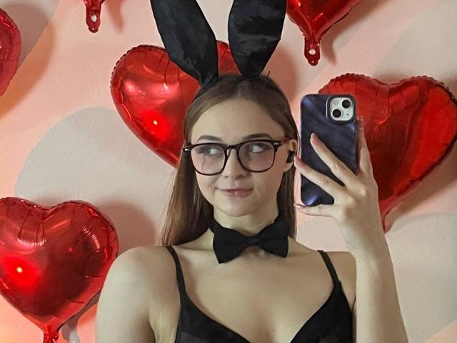 MayxFlower with glasses, black bunny ears, bowtie, and black bra, holding phone, smiling, red heart balloons in background, taking selfie, showing tits, cute, sexy.