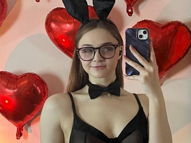 MayxFlower com óculos, orelhas de coelho pretas, laço borboleta e blusa preta transparente, segurando um telefone, sorrindo, balões em formato de coração vermelho, cabelo castanho longo, pele clara, seios médios, rosto fofo..
