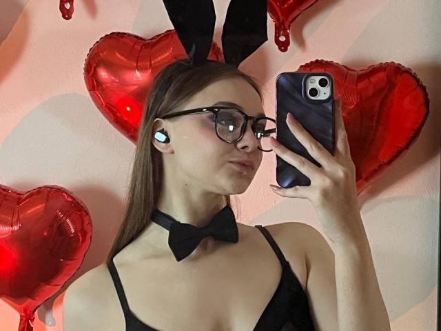 MayxFlower mit Brille, schwarzem Fliege und Hasenohren, die ein Selfie mit ihrem Handy macht, trägt ein schwarzes Oberteil, zeigt ihre Brüste, im Hintergrund sind rote Herzballons zu sehen..