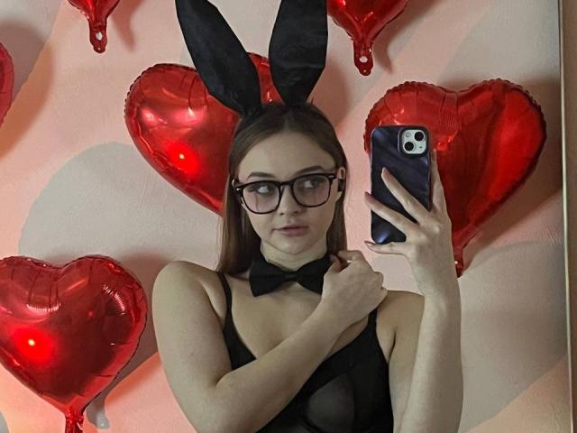 MayxFlower con gafas, orejas de conejo negras, lazo y una blusa negra transparente, sosteniendo un teléfono, globos en forma de corazón rojo en el fondo, reflejados en un espejo..