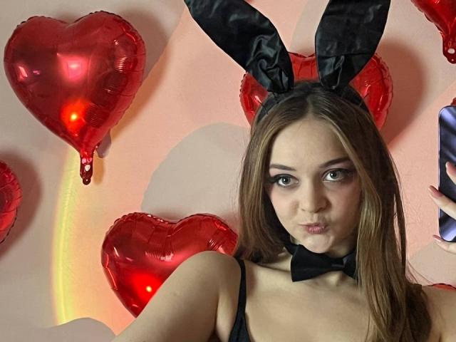 MayxFlower met lang bruin haar, zwarte konijnenoren en een strikje, met een frons, draagt een zwarte top, rode hartvormige ballonnen en heeft een telefoon aan de rechterkant..