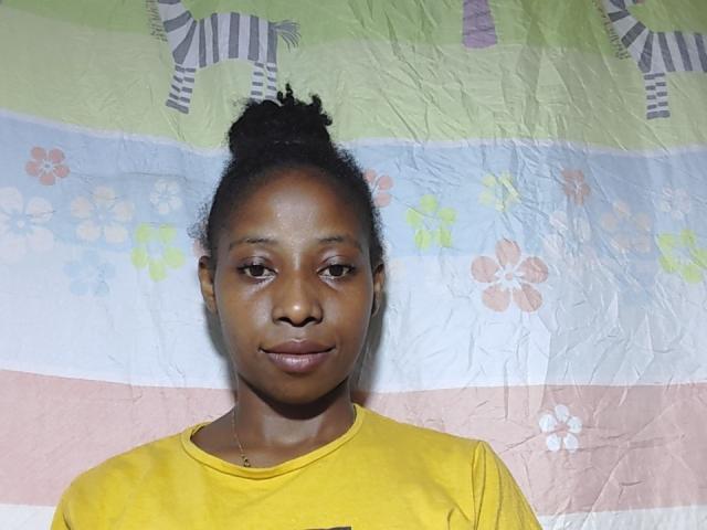 JeydaE has dark brown skin, a curly top bun, and wears a yellow shirt. Her small tits and subtle smile are visible. She stands in front of a colorful, creased fabric.