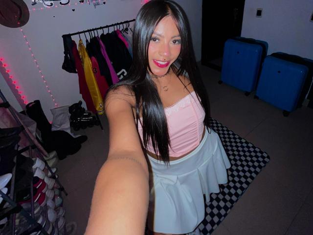 Amberv has long black hair, pink tube top, and white skirt, smiling at the camera with pink lipstick. She's holding out her arm for a selfie. Boobs, ass visible.