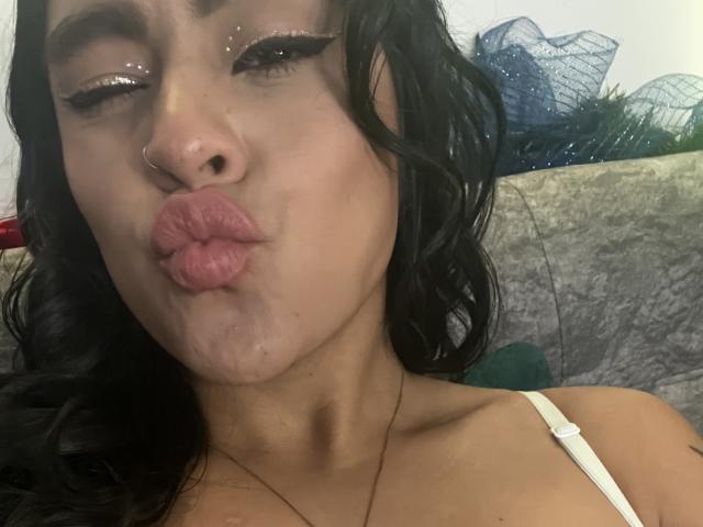 StellaBeckker with black wavy hair, wearing a white top, puffy pink lips, shiny eyelids, nose ring, and a gold necklace, making a puckered face.