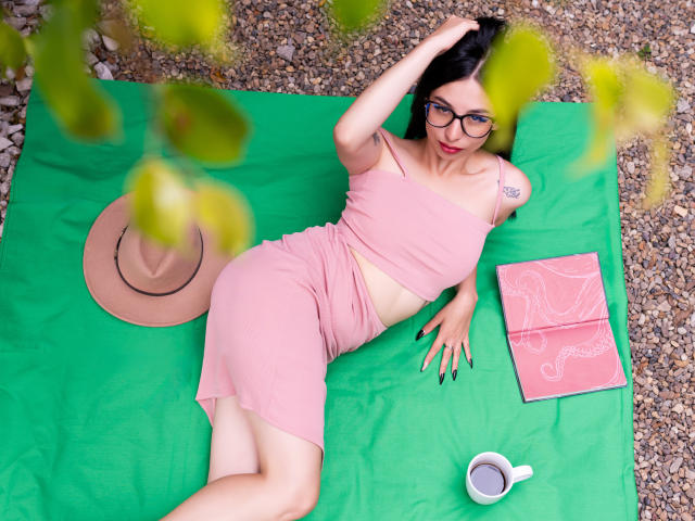 CamilleRios, with glasses, pink outfit, and black nails, lies on a green blanket, hand in hair, beside a hat, book, and cup, showing a tattoo on her right shoulder.