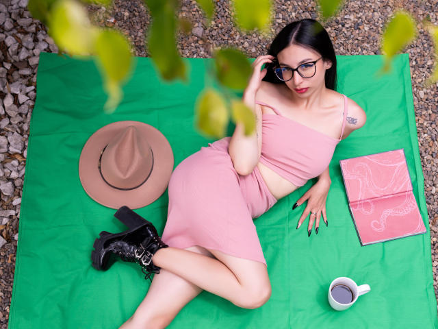 CamilleRios with black hair, glasses, pink outfit, black boots, lying on green blanket, hat, pink notebook, and white cup, showing ass and tattoo, in outdoor setting.