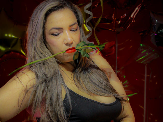 Mariehss, con cabello largo de color grisáceo-marrón, una camiseta negra, sosteniendo una rosa roja cerca de su boca, mirando hacia abajo. Sus senos son visibles, y tiene un tatuaje en su brazo derecho..