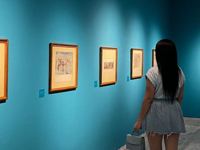 NovaDixon, with long black hair and a tattoo on her right arm, stands in front of four framed artworks on a blue wall, holding a white bag.