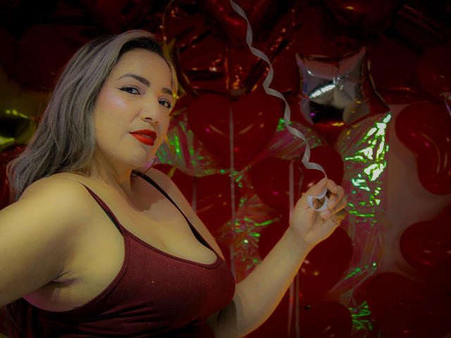 Mariehss, avec du rouge à lèvres, des cheveux gris et un débardeur rouge scintillant, tenant un ruban de curling blanc, montrant ses seins volumineux dans un décor coloré et festif..