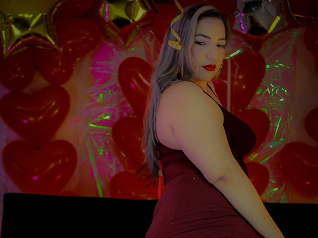 Mariehss with long gray hair, red lipstick, and yellow hair clip, wearing a tight red dress, showing her big ass and small tits, standing in front of red heart balloons.